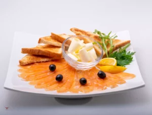 Assiette blanche élégante avec des tranches fines de saumon fumé disposées en éventail, accompagnées de toasts grillés, d’olives noires, de bâtonnets de fromage, de quartiers de citron et de feuilles de persil frais. Présentation raffinée idéale pour une entrée ou un apéritif maison.