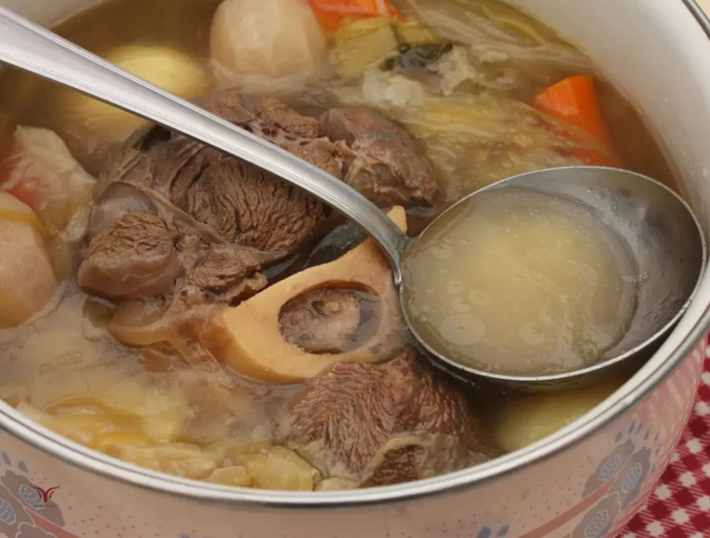 Cuisson du pot-au-feu traditionnel dans cocotte : viande de bœuf avec os à moelle et légumes mijotant dans bouillon doré, louche prélevant le bouillon parfumé